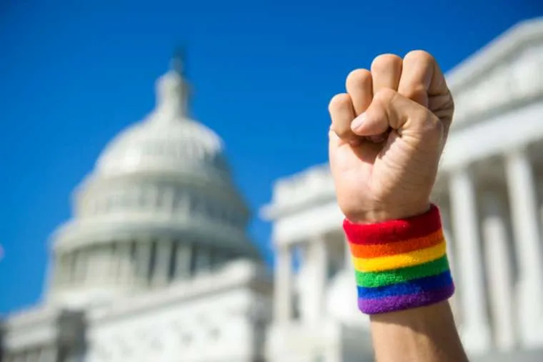 Hand wearing gay pride rainbow wristband making a power fist gesture in front of the US Capitol Building in Washington, DC. Via Shutterstock?w=200&h=150