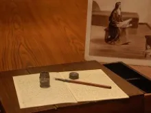 St. Therese of Lisieux's writing desk sits on display in front of a drawn image of the saint holding it in her lap. 