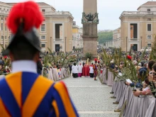Swiss Guards and faithful pilgrims holding olive branches line the processional route in St. Peter’s Square for Palm Sunday celebrations, April 13, 2025. The ancient Vatican obelisk stands at the center of the square as clergy process toward the basilica.