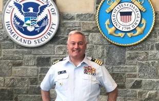 Father Daniel L. Mode, chaplain of the Coast Guard, stands in the large atrium of the Coast Guard headquarters in Washington, D.C., in front of a massive wall of flags and insignia of the Department of Homeland Security and the U.S. Coast Guard. Credit: Leslie Miller/Arlington Catholic Herald