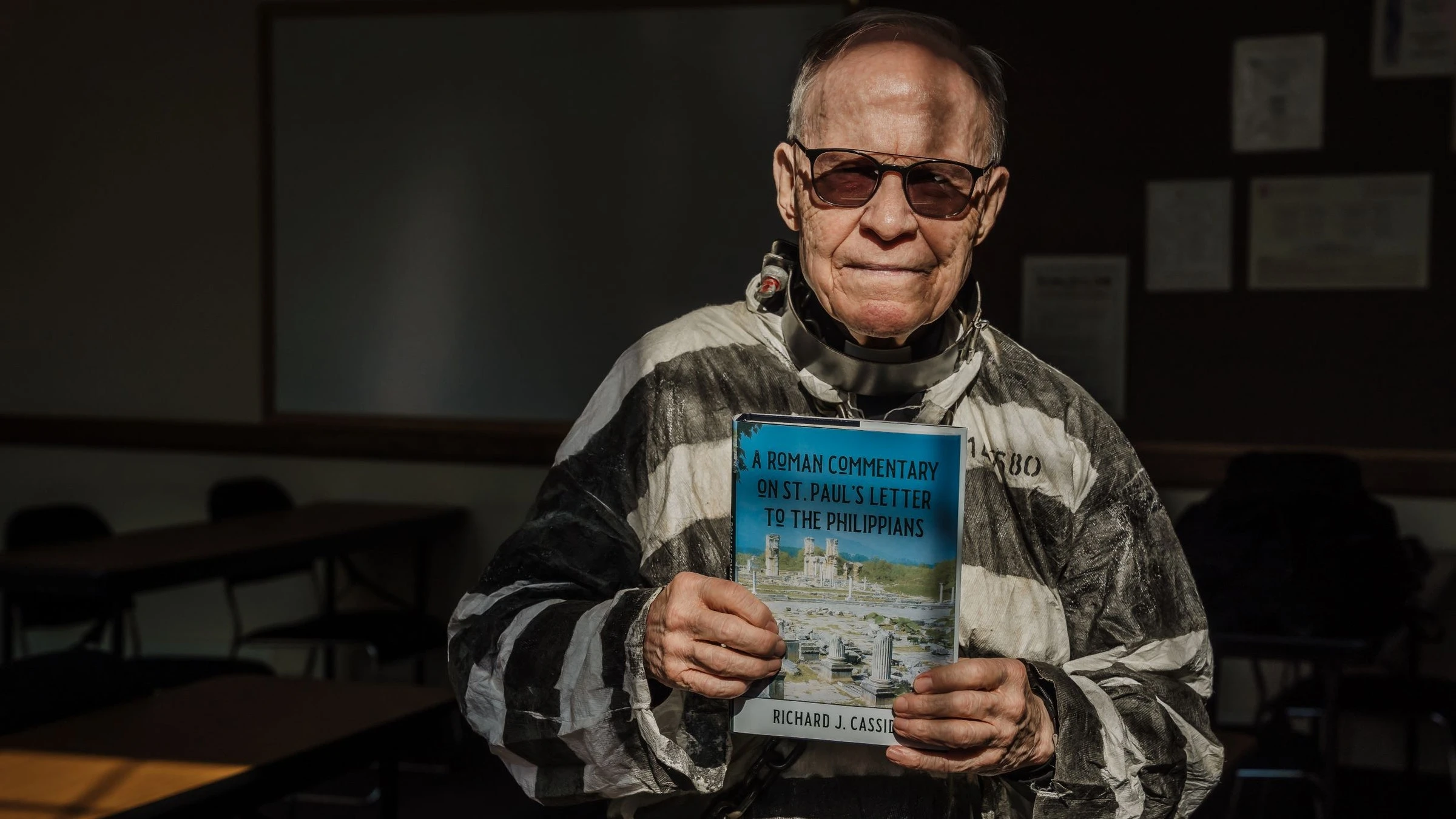 Fr. Richard Cassidy, professor of Sacred Scripture at Sacred Heart Major Seminary, dresses in Roman prisoner garb as he holds a copy of his newest book, "A Roman Commentary on St. Paul’s Letter to the Philippians." Fr. Cassidy's eighth scholarly work, the book explores the subversive nature of St. Paul's Letter to the Philippians, which the apostle wrote from behind bars in a Roman prison cell.?w=200&h=150